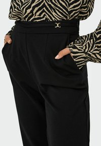 Donna che indossa pantaloni neri a vita alta con le mani nelle tasche e una maglietta a maniche lunghe con stampa zebrina infilata dentro.