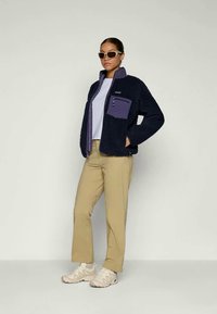Marinefarget fleecejakke med en lilla brystlomme, kombinert med beige bukser og kremfargede sneakers. Modellen har også på seg hvite solbriller.
