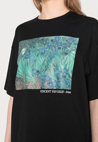 Czarny T-shirt z dużym nadrukiem graficznym "Irysy" Van Gogha w żywych odcieniach zieleni i błękitu; prosty okrągły dekolt, krótkie rękawy.