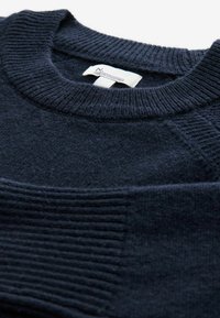 Mornarsko modra pletena sv sweater z rebrastim ovratnikom in teksturiranim vzorcem spredaj. Mehak, fin material z oznako "Premium Collection."