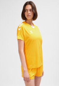 Gult sportset bestående av tröja och shorts i lättviktstyg, med V-ringning, korta ärmar och vita chevrondetaljer på axlarna.