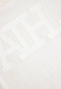 Koszulka w kolorze kremowym z uniesionym białym logo z napisem "ATHL" o teksturowanym wykończeniu. Miękka tkanina z widocznymi detalami szwów.