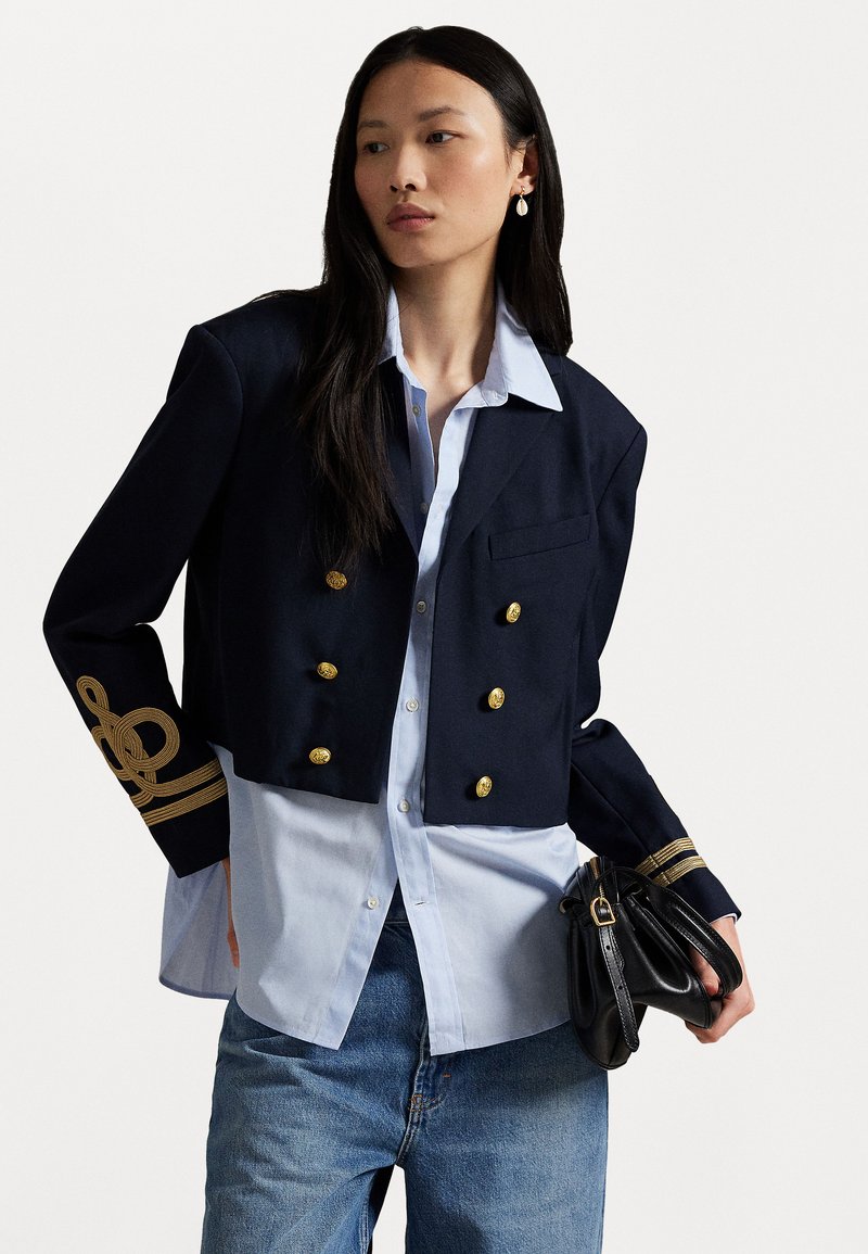 Navy cropped jacket in a wool blend with gold buttons and braided accents on sleeves, paired with a light blue shirt and black bag.