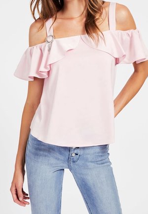 Kobieta w jasnoróżowej bluzce odkrywającej ramiona z falbankami i srebrną broszką, w połączeniu z jasnoniebieskimi dżinsami.