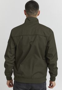 Maslinasto zelena bomber jakna s visokim ovratnikom, elastičnim manžetama i rubom. Izrađena od glatkog materijala s mat završnom obradom i minimalnim detaljima.