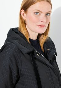 Černá vzorovaná bunda s kapucí, se zapínáním na zip a zlatými akcenty. Hladký povrch s elegantním, moderním designem.
