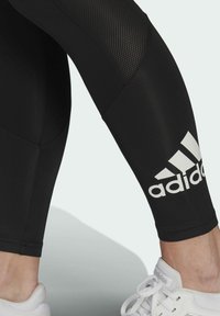 Czarne legginsy sportowe z siateczkowym wstawką i białym logo Adidas na łydce. W zestawie z białymi sneakersami. Gładki, elastyczny materiał.