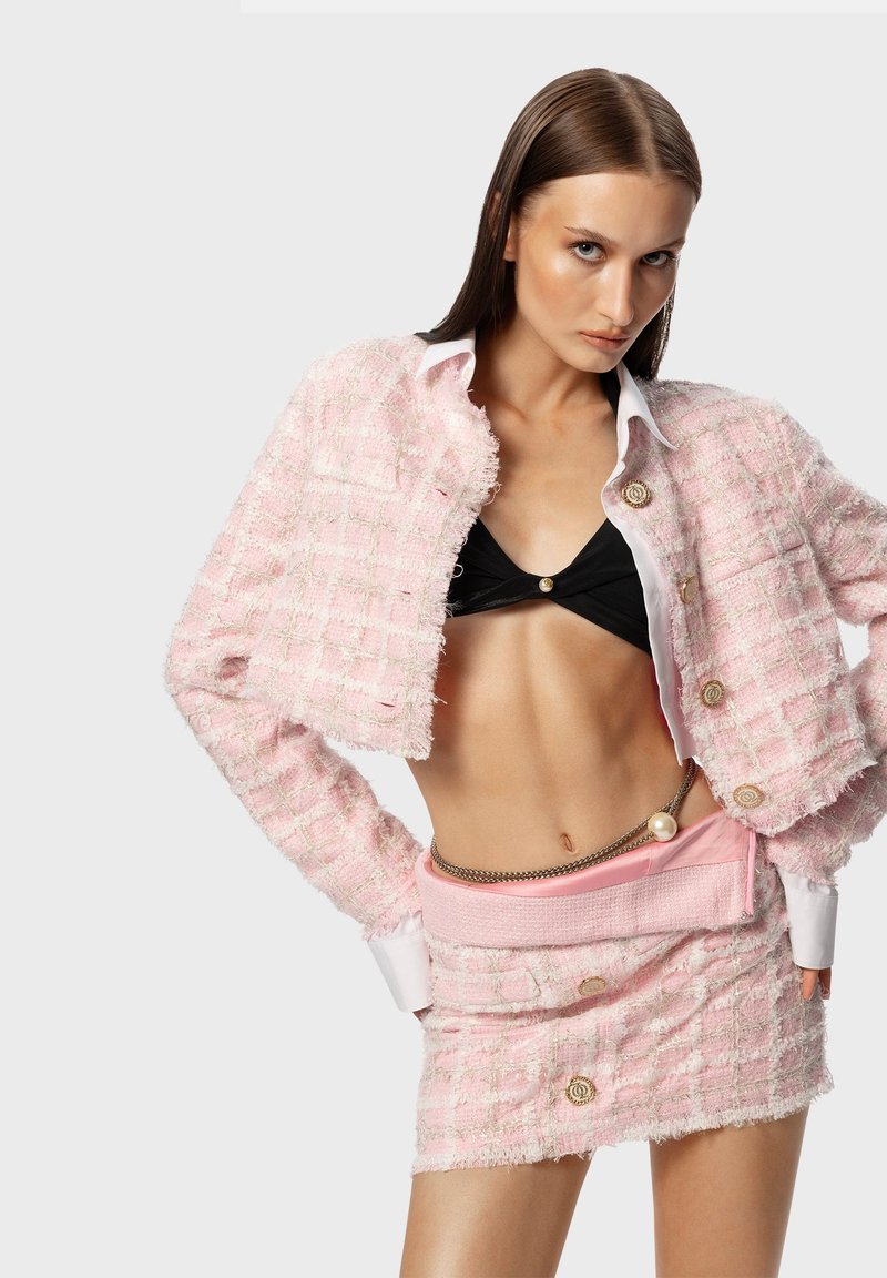Frau posiert in einer rosa Tweed-Jacke mit cropped Schnitt und passendem Minirock über einem schwarzen Bandeau-Top, ergänzt durch einen Kettengürtel und eine Perlenaccessoire.