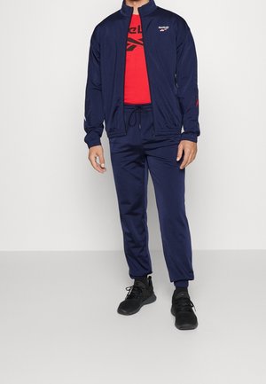 Mand iført navyblå Reebok træningsdragt med rød T-shirt med grafik og sorte sportssko, stående mod en ensartet lys baggrund.