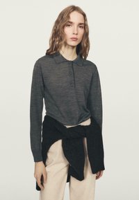 Koszulka polo z długim rękawem zapinana na guziki, wykonana z ciemnoszarej dzianiny, zestawiona z jasnobeżowymi spodniami i czarnym swetrem zawiązanym węzłem wokół talii.