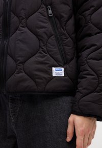 Black quilted jacket with zipper pockets and a white "HUGO" brand label on the front, worn over dark gray denim jeans.