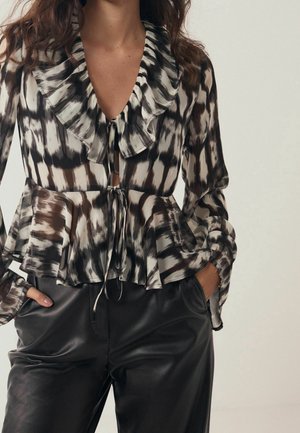 Mujer con una blusa de volantes con estampado en blanco y negro y cierres de lazo, combinada con pantalones de cuero negros, con las manos en los bolsillos.