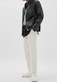 Veste en cuir noir de moto avec des accents matelassés aux épaules, associée à un t-shirt graphique noir et un pantalon cargo beige. Des baskets blanches complètent le look.