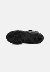 Černá gumová podrážka boty s texturovaným vzorem pro lepší přilnavost, obsahující hladkou část na patě a malou značkovou etiketu blízko kotníku.