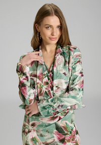 Ensemble blouse et jupe fleuri avec une base verte ornée de motifs de lys roses et blancs. Manches longues avec un décolleté drapé. Texture de tissu lisse.