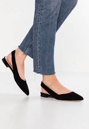 Persoon die zwarte suède slingback platte schoenen draagt met puntige neuzen en versleten blauwe spijkerbroek op een witte vloer.