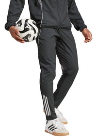 Schwarze sportliche Jogginghose mit seitlichem schwarzen Einsatz, weißen Streifen an den Unterschenkeln, strukturiertem Stoff, kombiniert mit einem Fußball und weißen Turnschuhen.