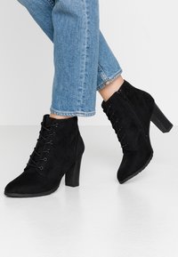 Botines de ante negro con tacón alto, que presentan detalles de cordones y cierre de cremallera, junto con una suela texturizada y diseño de punta afilada.