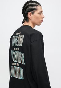 Schwarzer Pullover mit langen Ärmeln und mehrfarbigem Text auf der Rückseite. Das Material wirkt weich, mit einem lockeren Schnitt und Rundhalsausschnitt.