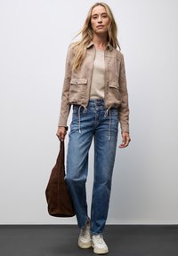 Tan suède cropped jack met trekkoordonderkant, beige top, lichtblauwe straight-leg jeans en witte sneakers. Bruine slouchy tas in de hand.