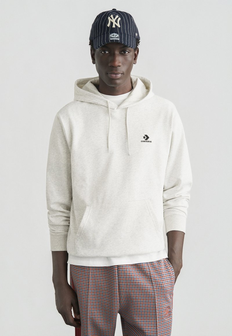 Sweat à capuche gris avec poche kangourou et logo Converse noir, associé à un pantalon à carreaux. Une casquette rayée noire et beige complète la tenue.