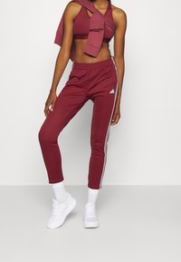 Burgunderfarbene Sporthose mit weißen Seitenstreifen und einem elastischen Bund, kombiniert mit einem passenden Sport-BH und weißen Sneakers.