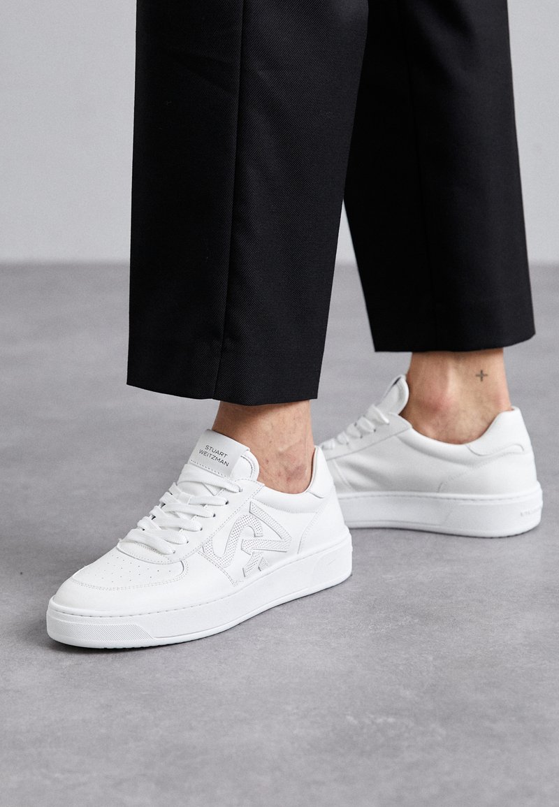 Sneakers bianchi con finitura in pelle liscia, dettagli perforati, lacci piatti e logo sulla linguetta. Abbinati a pantaloni neri a vita alta.