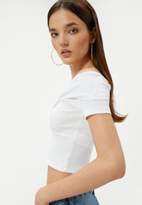 Hvid stribet crop top med korte ærmer, der har detaljeret draperet halsudskæring. Modellen bærer denimshorts og store øreringe i guld.