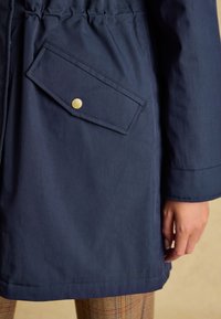 Cappotto blu navy con una tasca a patta diagonale chiusa da un bottone automatico dorato, indossato sopra pantaloni a quadri, con la mano destra visibile.