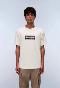 Beżowy bawełniany t-shirt z krótkimi rękawami, z czarną prostokątną grafiką na środku. Model nosi beżowe spodnie typu cargo.