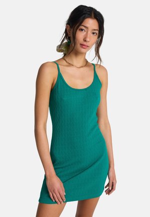 Junge Frau trägt ein eng anliegendes grünes ärmelloses Kleid mit dünnen Trägern und einer goldenen Kettenkette, posiert vor einem schlichten weißen Hintergrund.