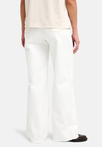 Λευκό τζιν καμπάνα από βαμβάκι, με άγρια τελείωμα, σχέδιο με πέντε τσέπες και μερικώς ορατό ανοιχτό μπεζ πουκάμισο από πάνω.