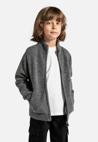 Szary sweter na zamek z prążkowaną fakturą wokół dekoltu i mankietów, noszony na białej koszuli, zestawiony z czarnymi spodniami typu cargo.