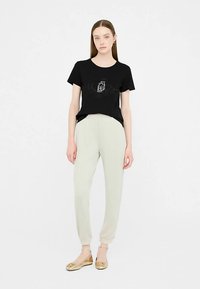 Zwart katoenen T-shirt met een grafisch ontwerp, gecombineerd met lichtgrijze taps toelopende broek. Model draagt gouden platte schoenen. Minimalistische casual outfit.