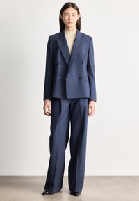 Žena stojí, má na sobě tmavě modrý dvouřadý blejzr a odpovídající široké kalhoty, béžový rolák a černé boty na neutrálním pozadí.