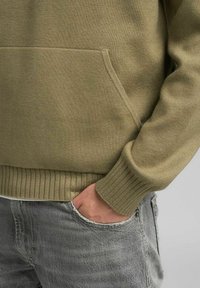 Maglione verde oliva con polsini e orlo a coste; presenta una tasca frontale e un tessuto morbido e strutturato. Indossato sopra jeans grigi con effetto consumato.