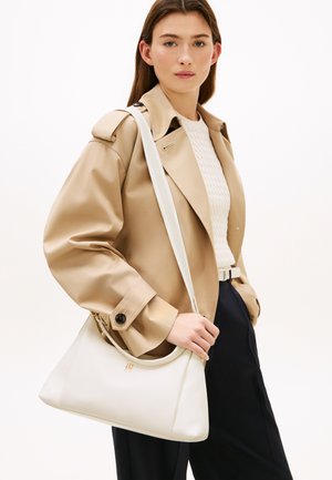 Femme portant un trench-coat beige et un haut en maille blanc, tenant un sac à bandoulière en cuir blanc avec un logo doré, sur un fond blanc uni.