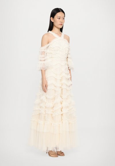 Off-shoulder crème jurk met gelaagde ruches en doorschijnende mouwen. Gemaakt van textuurstof met een vloeiende rok. Sandalen maken de look compleet.