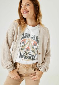 Licht beige gebreide cardigan met een textuurpatroon, gedragen over een witte graphic t-shirt met kleurrijke bloemenontwerpen, gecombineerd met beige broeken.