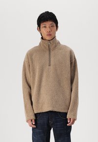 Beige fleecetrui met een hoge kraag en een kwart-ritssluiting. Zacht, gestructureerd materiaal met een relaxte pasvorm en lange mouwen.