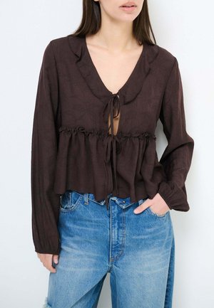 Donna che indossa una blusa a maniche lunghe marrone scuro con chiusure a fiocco e jeans a vita alta azzurri, mano in tasca, su sfondo bianco.