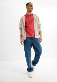 Felpa beige con zip sopra una maglietta polo rossa, pantaloni jeans blu larghi e sneakers bianche. Linee pulite e vestibilità casual.