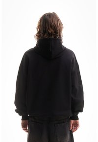 Personne avec des cheveux bruns ondulés portant un hoodie noir et un pantalon sombre, se tenant de dos sur un fond blanc.