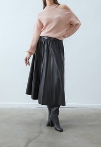 Pull rose clair côtelé avec un design épaules dénudées, associé à une jupe midi plissée noire et des bottes grises à talons montant jusqu'aux genoux.