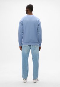 Sudadera azul claro con cuello redondo y puños acanalados, combinada con jeans de mezclilla azul claro. Modelo de espaldas a la cámara.