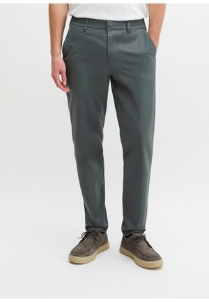 Mann trägt dunkelgraue Freizeithose und braune Schnürschuhe, steht mit einer Hand in der Tasche vor einem einfarbigen Hintergrund.