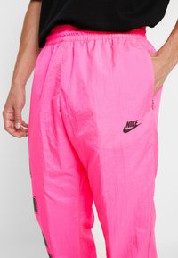 Person wearing bright pink Nike track pants with elastic waistband and black logo, one hand in pocket and the other relaxed by side.