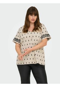 Beige blúz fekete geometrikus mintákkal, rövid ujjakkal és V-nyakkal. Az ujjakon hímzett díszítések találhatók, valamint gombos záródással rendelkezik.