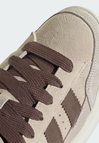Beige suède sneaker met bruine geweven veters, met drie bruine strepen, een gestructureerde neuskap en een witte rubberen zool.