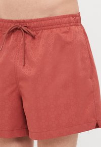 Røde svømshorts lavet af letvægtsmateriale, med et struktureret geometrisk mønster, elastisk talje med snoretræk til justering.